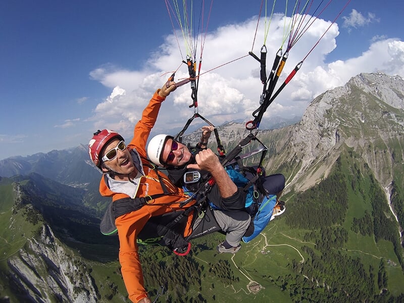 Parapente en tandem au-dessus d'un paysage montagneux impressionnant, avec des vallées verdoyantes en contrebas. L'instructeur, en combinaison orange, et le passager, en veste bleue, lèvent les bras e
