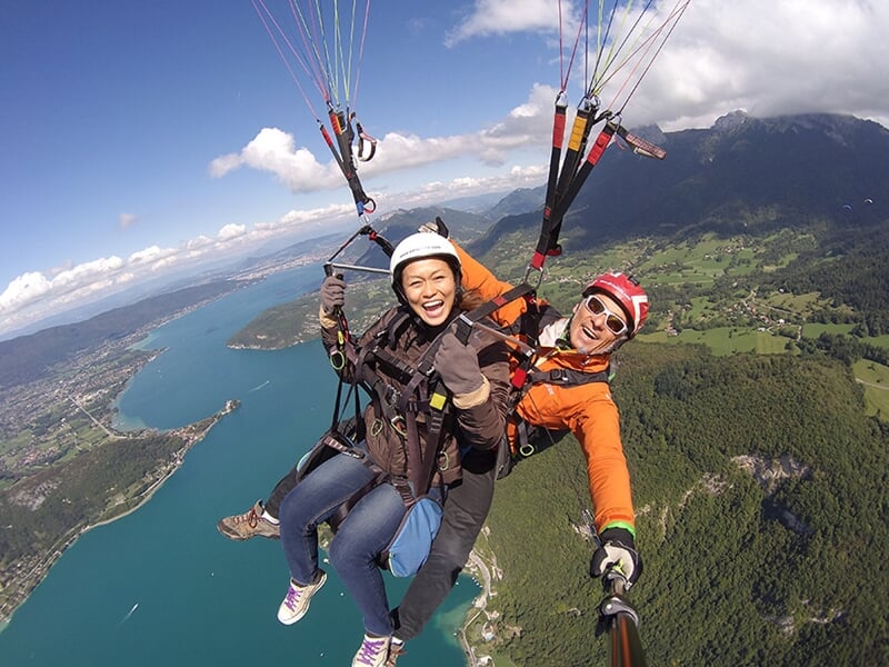 Parapente en tandem au-dessus d'un grand lac bleu, encadré par des montagnes et des forêts. L'instructeur, en combinaison orange, et le passager asiatique, vêtue de noir et de marron, expriment leur j