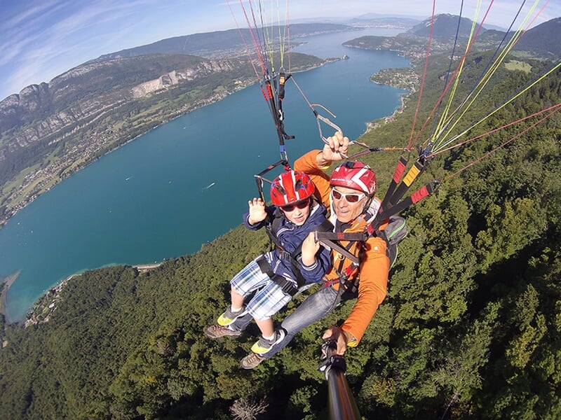Vue aérienne en parapente montrant un instructeur et un passager en tandem, survolant un paysage de lac entouré de montagnes verdoyantes. L'instructeur porte une combinaison orange et le passager une