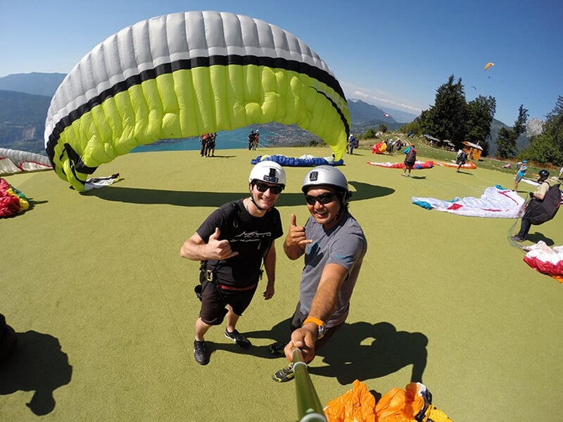 Deux parapentistes souriants équipés de casques et de harnais, font un signe de la main sur un site de décollage avec un parapente noir et vert en toile de fond. Ils sont entourés d'autres parapentes