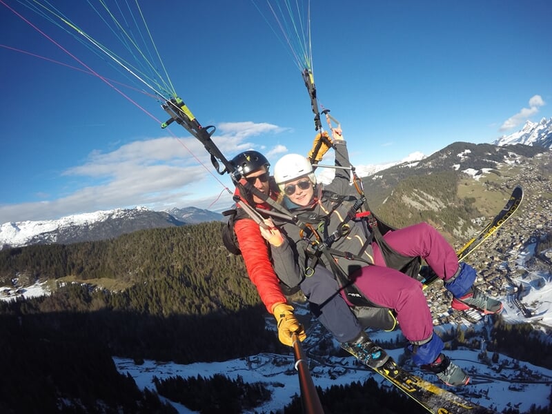 Parapente avec deux personnes en tandem survolant un paysage enneigé et boisé en montagne