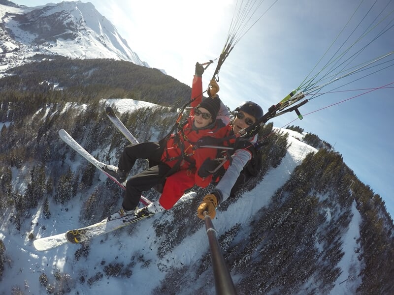 Parapente avec deux personnes en tandem survolant une vallée enneigée et des forêts