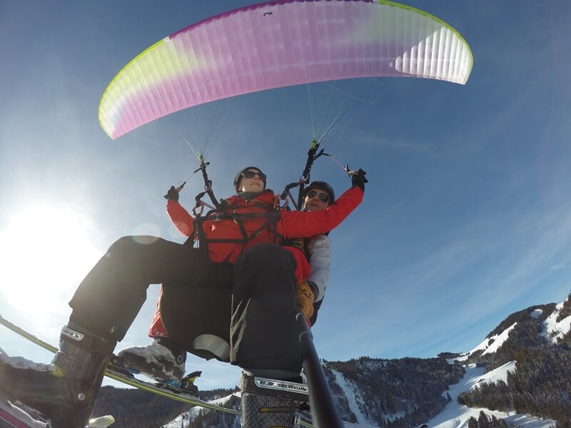 Parapente en tandem avec skieurs volant au-dessus d'un paysage montagneux enneigé