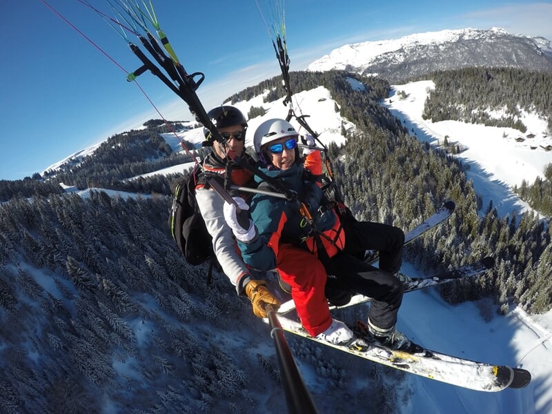 Parapente avec deux personnes en tandem survolant un paysage montagneux enneigé et boisé