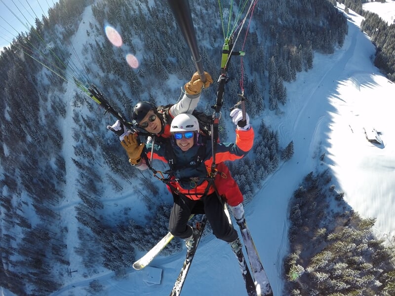 Parapente en tandem avec deux skieurs volant au-dessus d'une vallée enneigée et boisée