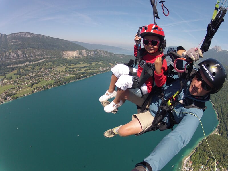 Parapente en tandem au-dessus d'un grand lac turquoise, avec un enfant en tenue rouge et casque assorti exprimant sa joie par un signe de la main et un instructeur