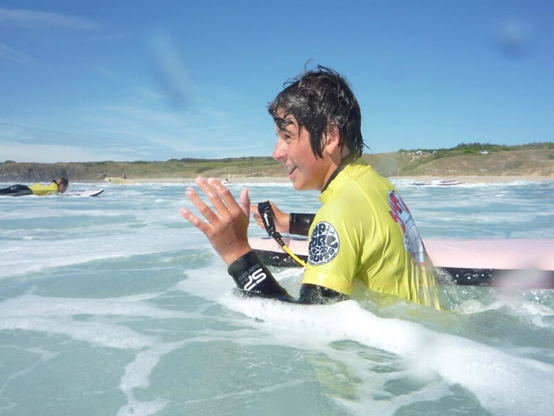 Jeune surfeur souriant dans l'eau avec une combinaison jaune, prêt à attraper une vague sur la plage de Crozon.