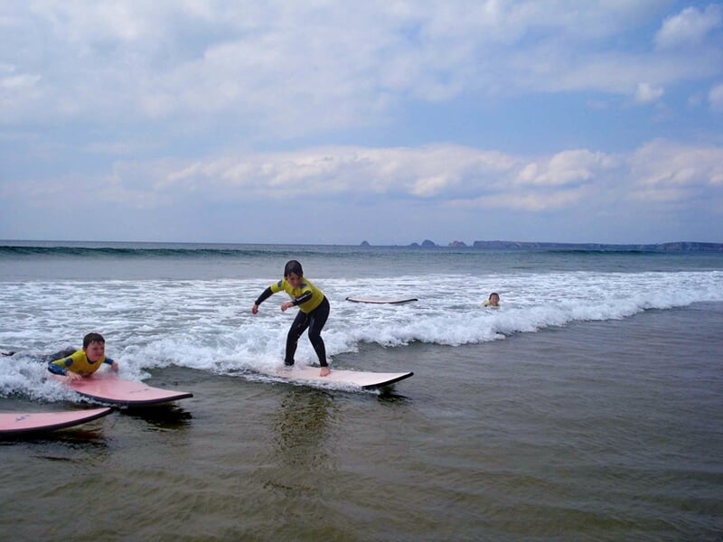 Deux surfeurs en combinaison jaune sur leurs planches, l'un debout et l'autre allongé, surfant sur une petite vague à Crozon.