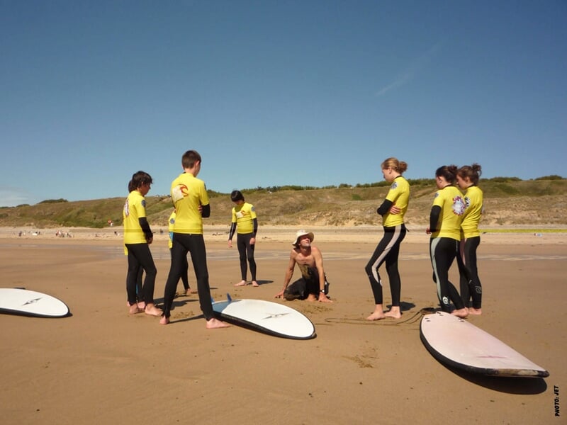 Groupe de surfeurs en combinaison jaune écoutant un instructeur assis sur le sable avec leurs planches sur la plage de Crozon.