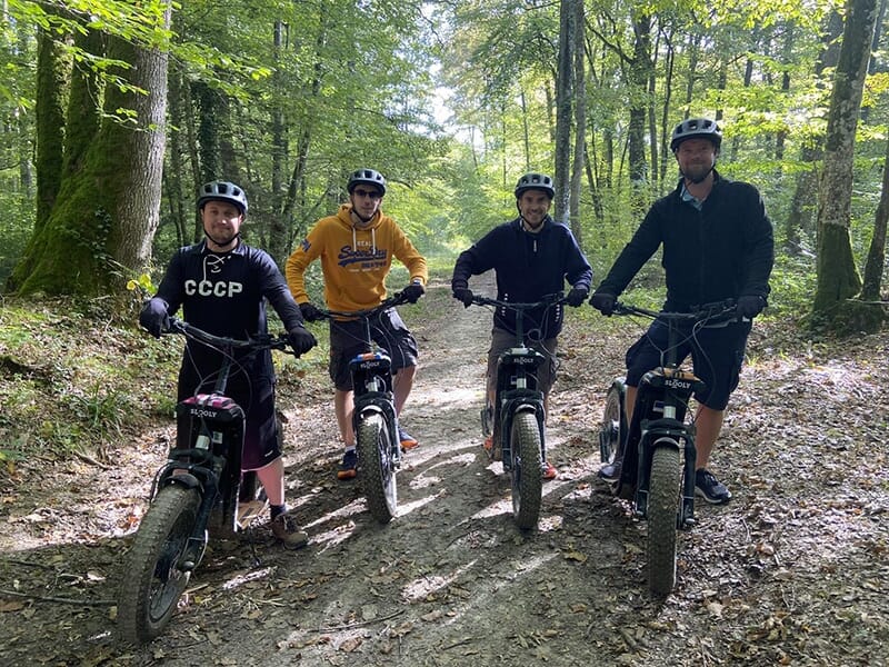 Quatre hommes en tenue de sport portant un casque et des gants posent pour la photo au guidon d'une trottinette électrique tout-terrain dans les sous-bois