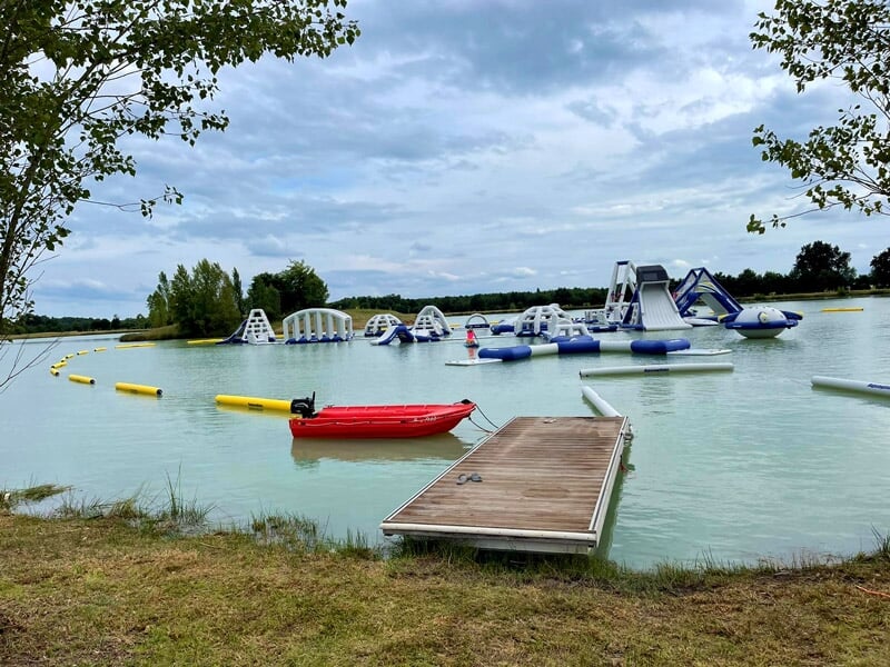 Vue d'un parc aquatique sur un lac avec structures gonflables, ponton en bois et ciel nuageux en arrière-plan.