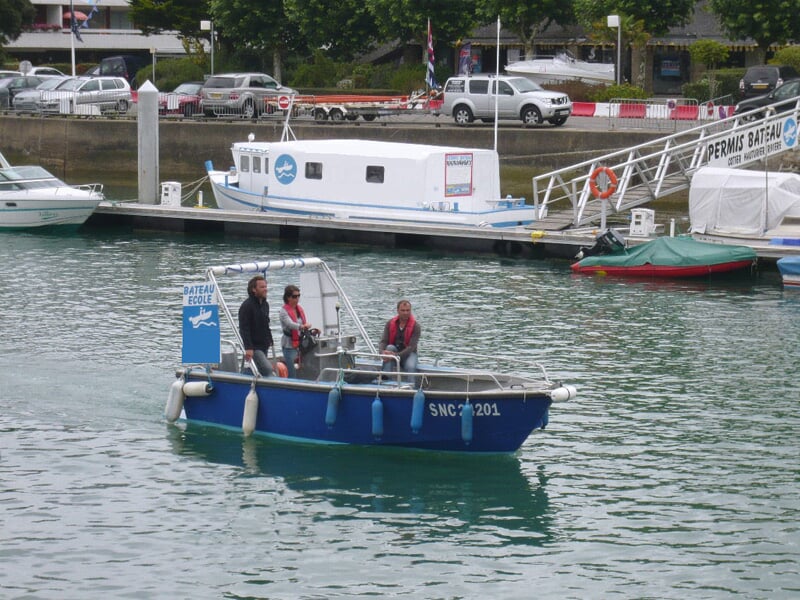 Trois personnes sur un petit bateau bleu naviguant dans un port avec des voitures et un autre bateau en arrière-plan.