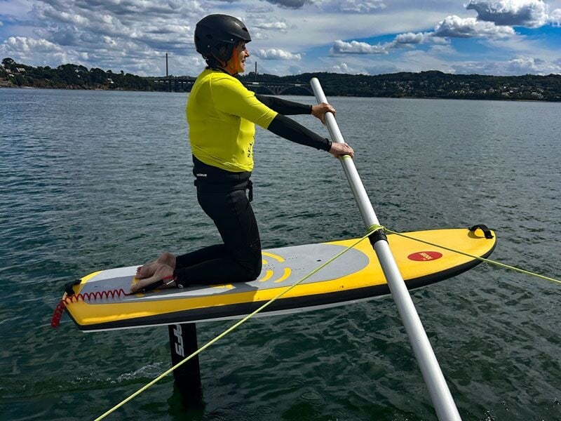Personne en tenue jaune et casque noir sur une planche de wingfoil posée sur l'eau calme d'un lac ou mer.