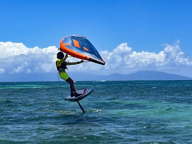 Sportif en combinaison jaune et casque noir pratiquant le wingfoil sur une mer turquoise sous un ciel bleu clair.