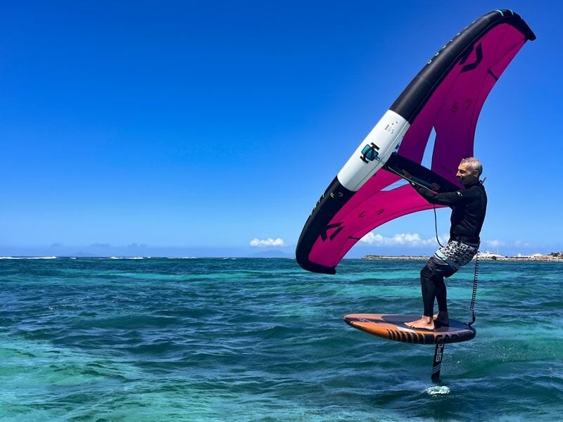 Homme en short noir et haut noir fait du wingfoil avec une voile rose sur une mer turquoise sous un ciel bleu.