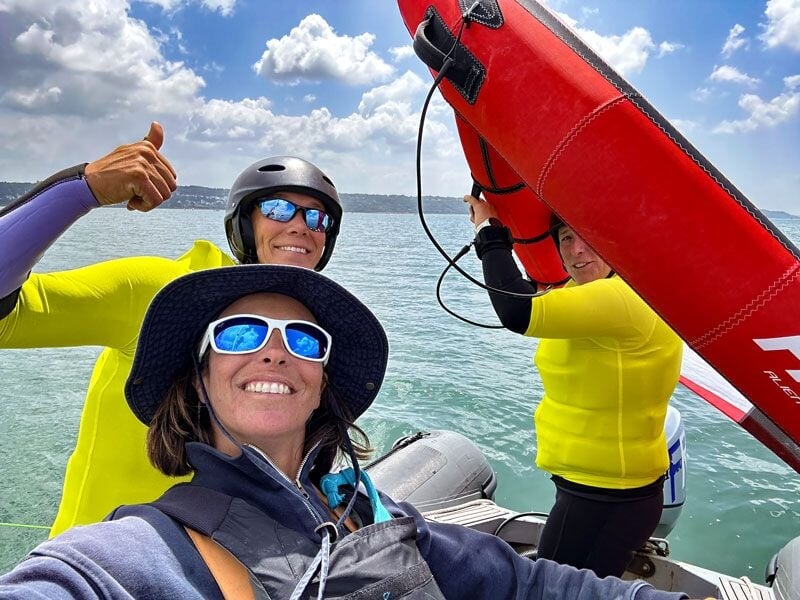 Trois personnes souriantes en tenue de sport jaune et lunettes de soleil sur un bateau gonflable au bord de l'eau.