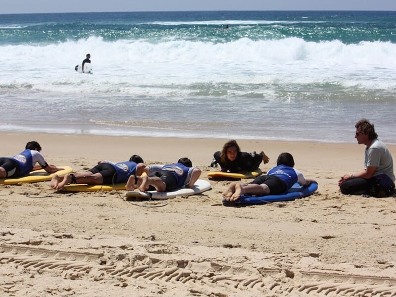 Groupe de débutants allongés sur des planches de surf sur la plage, un moniteur les observe attentivement.