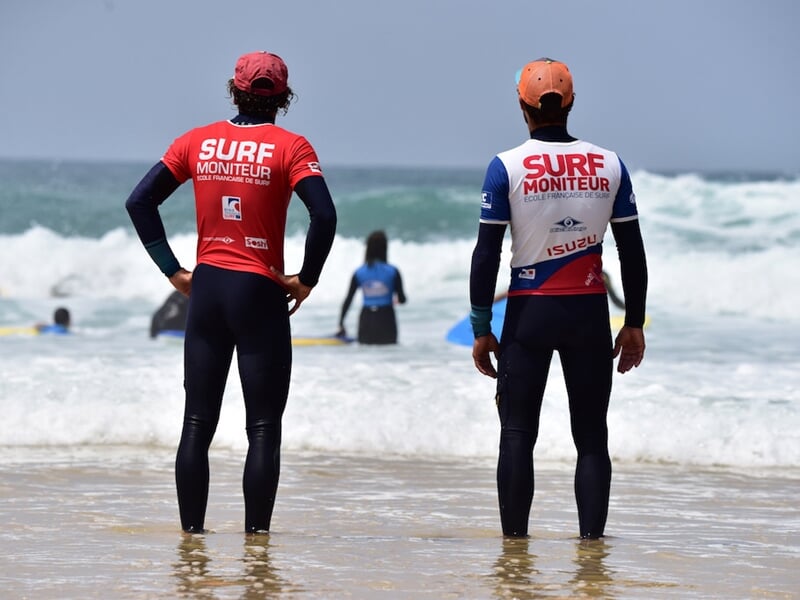 Deux moniteurs de surf en combinaison noire et tee-shirts rouges et blancs regardent la mer agitée à Seignosse.