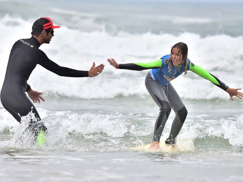 Un moniteur aide une jeune fille en combinaison bleue à tenir l'équilibre sur une planche de surf dans l'eau.