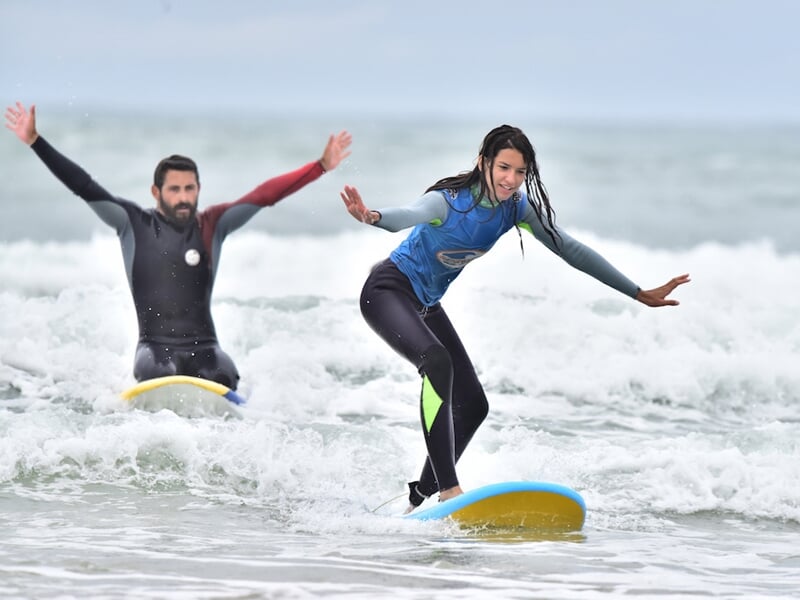 Un homme et une jeune fille surfent sur des petites vagues, tous deux en combinaison, souriants et concentrés.