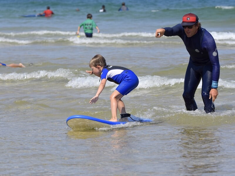 Un enfant en combinaison bleue apprend à surfer avec un moniteur qui le guide dans l'eau peu profonde.