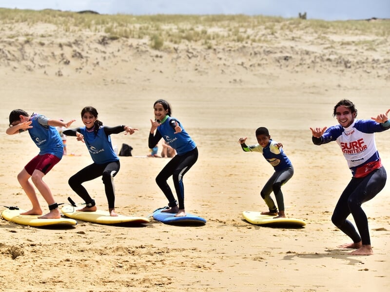 Groupe d'enfants en combinaison bleue et moniteur en tee-shirt rouge font une pose de surf sur le sable.