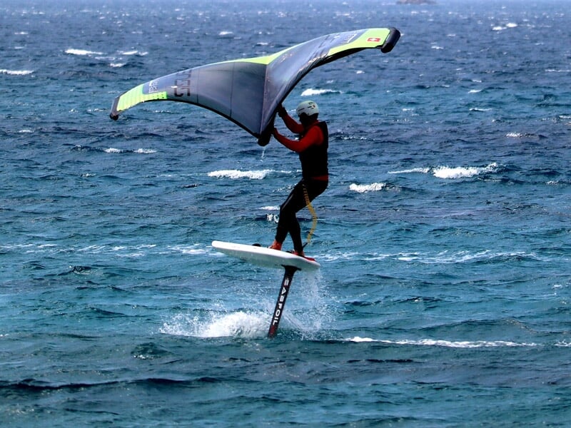 Surfeur en combinaison rouge et noir maîtrisant le wingfoil, tenant une aile verte et grise au-dessus des vagues, mer agitée et ciel bleu lumineux