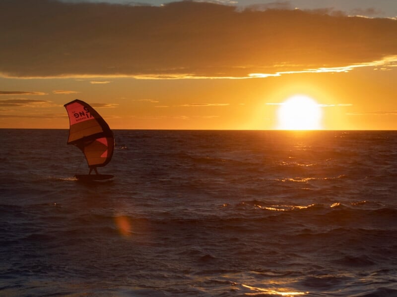 Navigateur solitaire en wingfoil à l’horizon, silhouetté par un soleil couchant orange vif, mer légèrement agitée et ciel nuageux