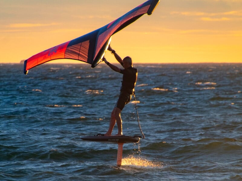 Pratiquant de wingfoil glissant sur la mer au coucher du soleil, aile bleue et rouge tendue dans le vent, lumière dorée illuminant l’eau