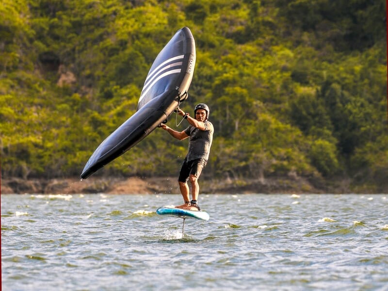 Homme en équilibre sur une planche de wingfoil, utilisant une aile noire avec rayures blanches, naviguant sur une eau calme bordée de collines verdoyantes