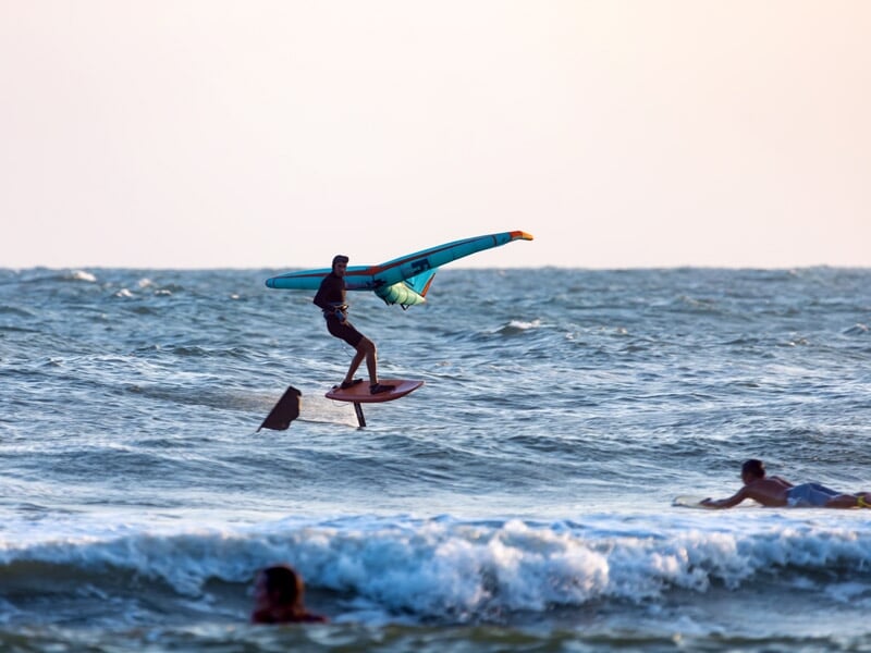 Surfeur pratiquant le wingfoil, debout sur une planche équipée d'un foil, tenant une aile turquoise et orange au-dessus des vagues en mer, sous un ciel clair
