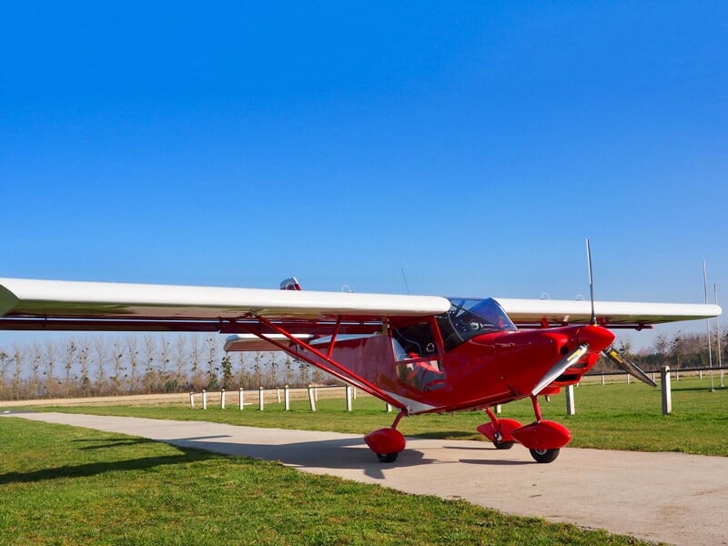 Petit avion ultraléger rouge stationné sur une piste en béton dans un champ vert sous un ciel bleu clair