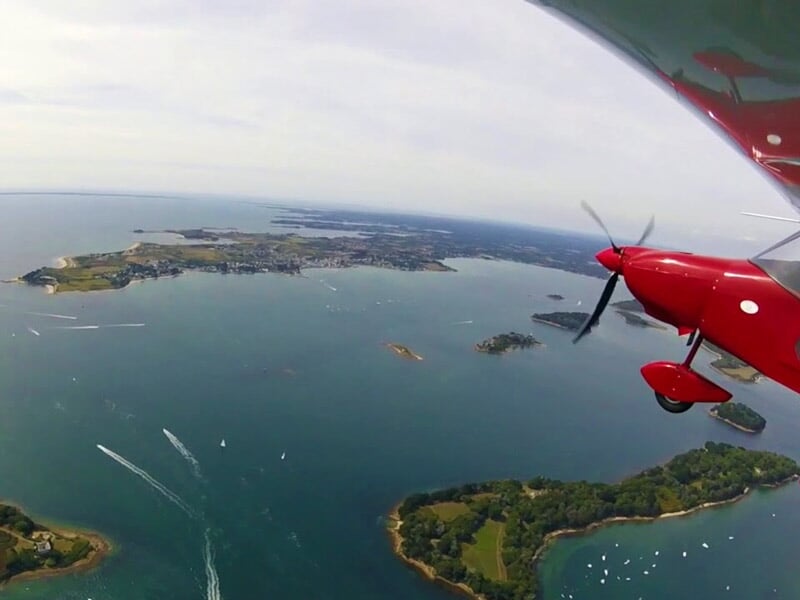 Vue aérienne prise depuis un petit avion rouge survolant une côte avec plusieurs îles et bateaux sur une mer calme.