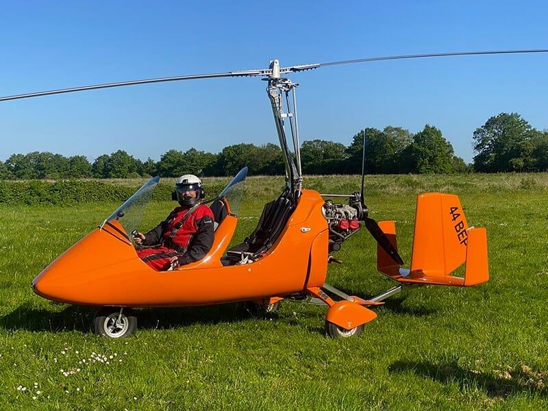 Pilote en combinaison rouge et casque dans un autogire orange sous un ciel bleu dégagé.