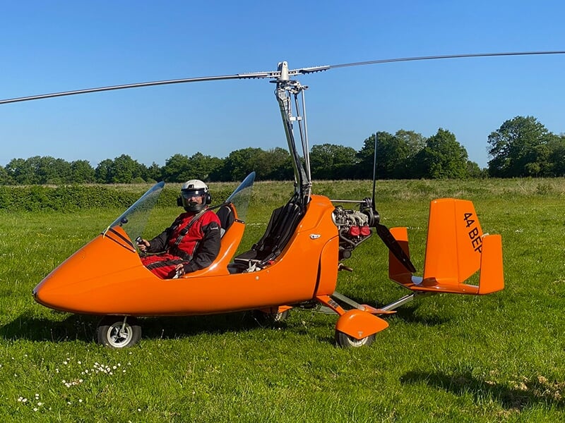 Pilote en combinaison rouge et casque dans un autogire orange sous un ciel bleu dégagé.