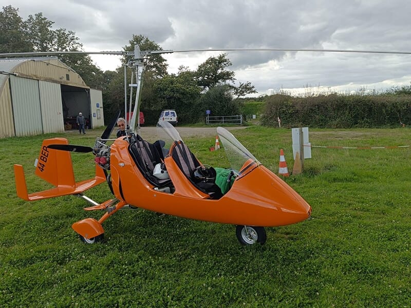 Autogire orange vide garé devant un hangar sous un ciel nuageux, avec matériel à proximité.