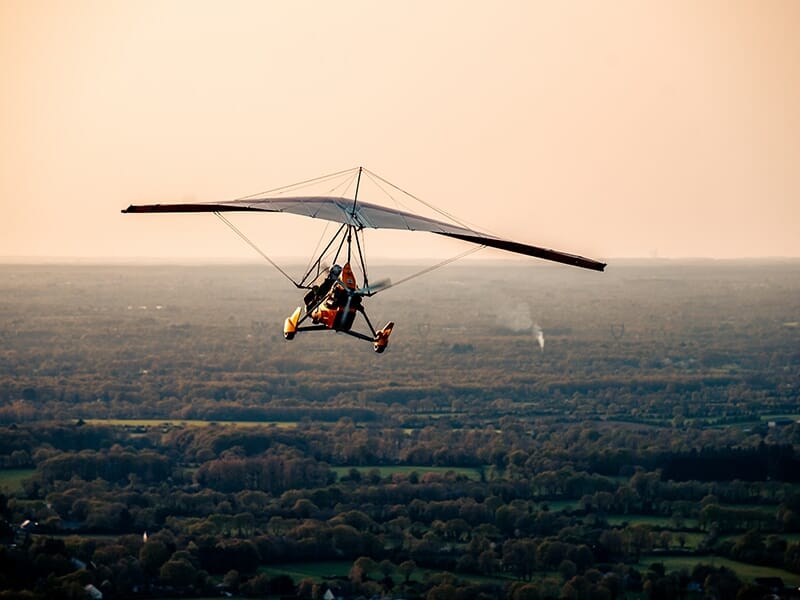 ULM pendulaire évoluant au-dessus d’un paysage vert entre forêts et rivières.