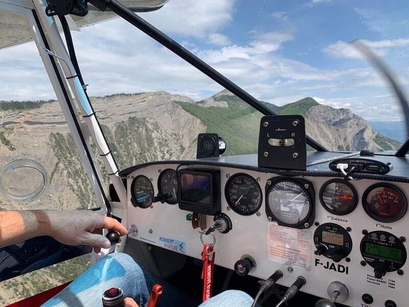 Vue depuis le cockpit d'un avion ULM avec un pilote aux commandes survolant un paysage montagneux sous un ciel partiellement nuageux.