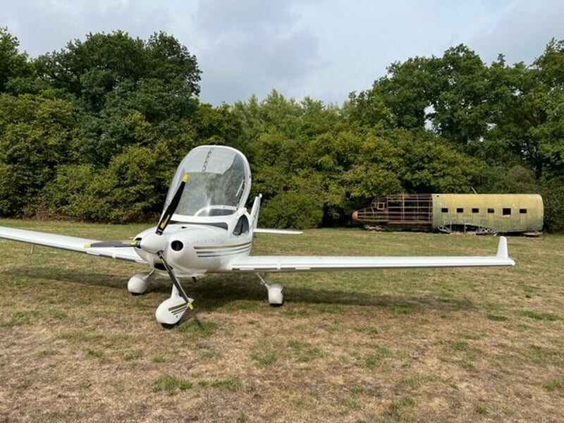 Petit avion multiaxes Dynamic blanc avec hélice sur un terrain herbeux, entouré d'arbres verts et d'une vieille carcasse d'avion rouillée.
