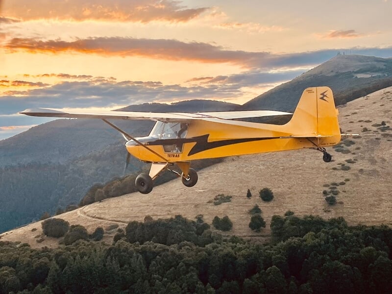 Avion ULM jaune volant au-dessus d'une colline boisée au coucher du soleil avec ciel aux teintes orangées.