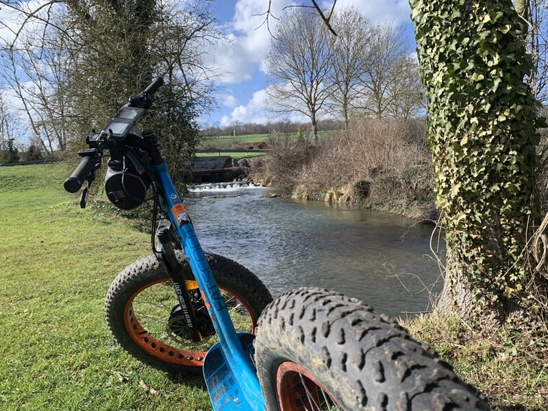 Trottinette électrique bleue avec pneus larges posée sur l'herbe près d'une rivière sous un ciel partiellement nuageux.
