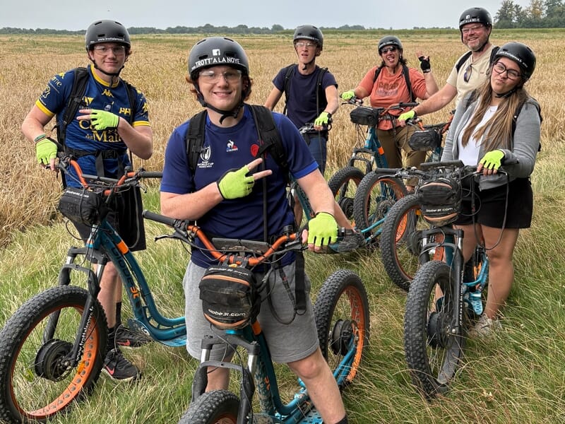 Groupe de six personnes souriantes avec casques et gants verts posant avec leurs trottinettes électriques en pleine nature.
