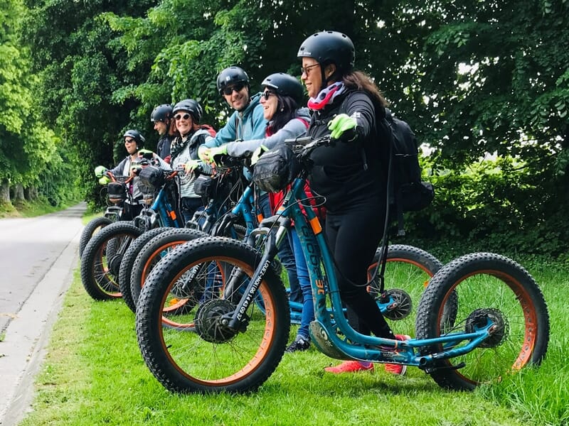 Groupe de personnes équipées de casques et trottinettes électriques alignées sur un chemin herbeux près d'une route.