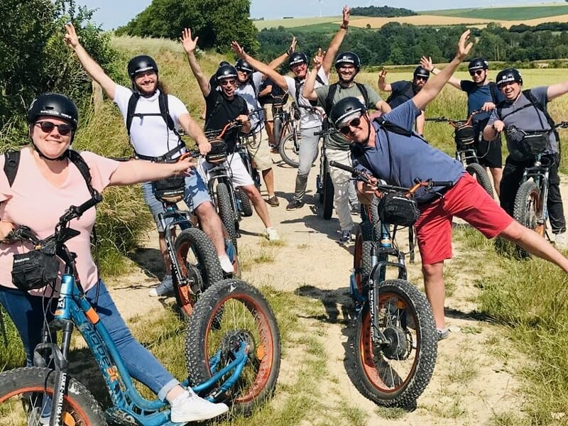 Groupe joyeux de neuf personnes avec trottinettes électriques posant sur un chemin ensoleillé dans un paysage champêtre.