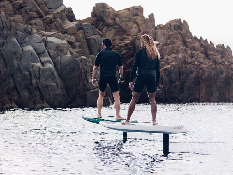 Deux personnes en combinaison de surf pratiquent le surf électrique sur une planche au-dessus de l'eau près des rochers