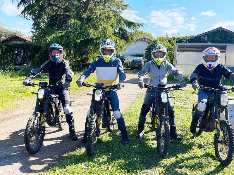 Quatre personnes équipées de casques et protections, posant sur des trottinettes électriques tout-terrain sur un chemin ensoleillé.