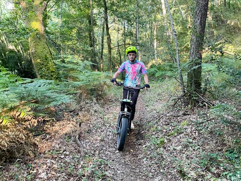 Jeune homme avec casque vert conduisant une trottinette électrique sur un sentier forestier entouré d'arbres et de fougères.