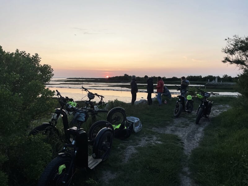 Groupe de personnes avec plusieurs trottinettes électriques garées près d'un chemin au bord de l'eau au coucher du soleil.