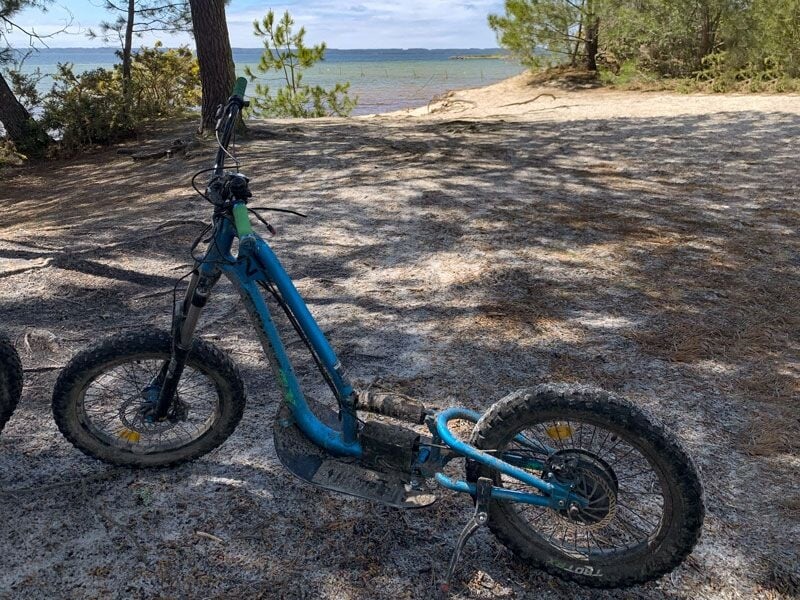 Trottinette électrique bleue tout-terrain garée sur un sol sablonneux près d'un lac avec arbres et ciel bleu en arrière-plan
