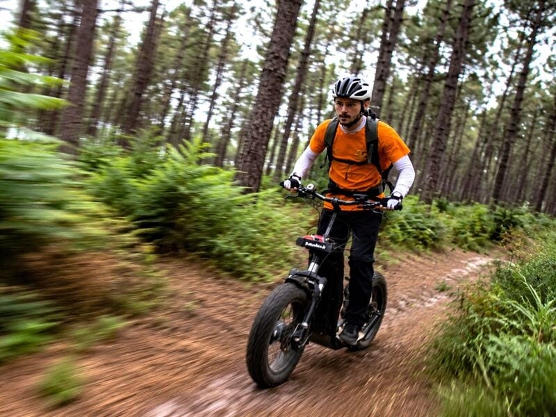 Homme en tenue orange et casque roulant sur une trottinette électrique tout-terrain dans une forêt dense et verdoyante.